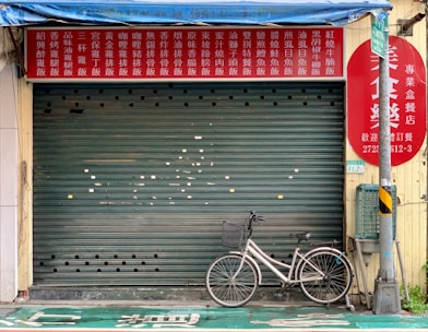 A closed shop with a red and yellow signboard featuring Chinese characters above a green rolling shutter. A bicycle is parked in front of the shop next to a utility pole.