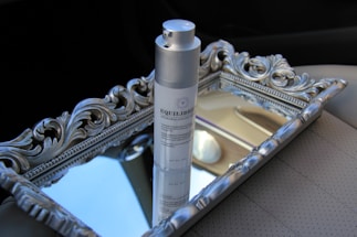 A bottle of skincare product labeled 'EQUILIBRIUM' stands on an ornate silver tray with decorative carvings, positioned on a car seat. The tray reflects parts of the interior, creating a luxurious and sophisticated arrangement.