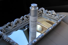 A bottle of skincare product labeled 'EQUILIBRIUM' stands on an ornate silver tray with decorative carvings, positioned on a car seat. The tray reflects parts of the interior, creating a luxurious and sophisticated arrangement.