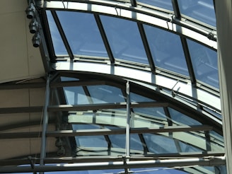 An architectural structure featuring a series of curved glass panels supported by metal frames. The glass provides an expansive view of the sky and allows natural light to enter. The metal beams intersect at various angles, indicating a complex, modern design.