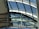 An architectural structure featuring a series of curved glass panels supported by metal frames. The glass provides an expansive view of the sky and allows natural light to enter. The metal beams intersect at various angles, indicating a complex, modern design.