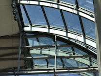 An architectural structure featuring a series of curved glass panels supported by metal frames. The glass provides an expansive view of the sky and allows natural light to enter. The metal beams intersect at various angles, indicating a complex, modern design.