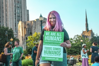 A person with purple hair is holding a green sign that reads 'We Are All Humans' and 'Todos Somos Humanos'. The scene is outdoors with a historical building and a spire in the background. Several other people are around, some holding signs, suggesting a public gathering or protest.