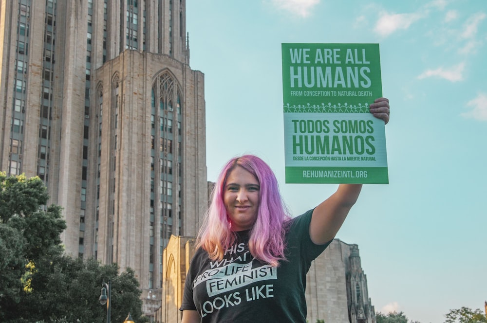 woman wearing black and white printed t-shirt standing while holding banner with we are all human text