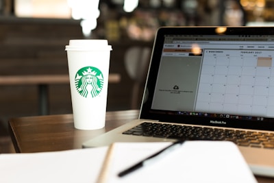 A coffee shop setting featuring a disposable coffee cup with a recognizable logo next to an open laptop displaying a calendar. The environment suggests a workspace or casual meeting place with a focus on productivity. A notebook is partially visible in the foreground, emphasizing the theme of planning or organization.