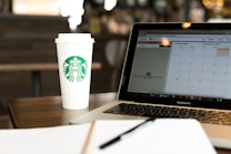A coffee shop setting featuring a disposable coffee cup with a recognizable logo next to an open laptop displaying a calendar. The environment suggests a workspace or casual meeting place with a focus on productivity. A notebook is partially visible in the foreground, emphasizing the theme of planning or organization.