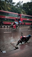 A street scene in rainy weather featuring a red double-decker bus labeled 'BRTC' parked by the roadside. A cyclist pedals a rickshaw, covered with a vibrant, colorful canopy to protect from the rain, along the wet street. A motorbike is parked on the wet pavement, and lush green palm trees form a backdrop.