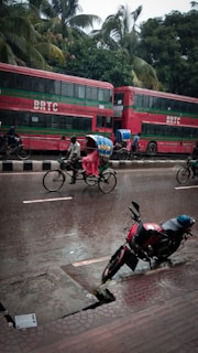 A street scene in rainy weather featuring a red double-decker bus labeled 'BRTC' parked by the roadside. A cyclist pedals a rickshaw, covered with a vibrant, colorful canopy to protect from the rain, along the wet street. A motorbike is parked on the wet pavement, and lush green palm trees form a backdrop.