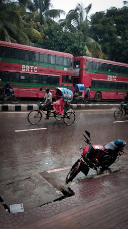 A street scene in rainy weather featuring a red double-decker bus labeled 'BRTC' parked by the roadside. A cyclist pedals a rickshaw, covered with a vibrant, colorful canopy to protect from the rain, along the wet street. A motorbike is parked on the wet pavement, and lush green palm trees form a backdrop.