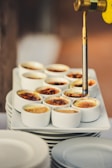 A culinary torch caramelizes the tops of multiple crème brûlées arranged neatly on a stack of plates. The custard desserts display a range of browned sugar textures, from light golden to dark amber, indicating the varying degrees of torched sugar crusts.