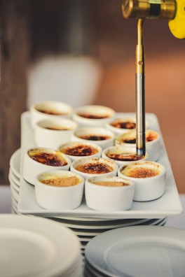 A culinary torch caramelizes the tops of multiple crème brûlées arranged neatly on a stack of plates. The custard desserts display a range of browned sugar textures, from light golden to dark amber, indicating the varying degrees of torched sugar crusts.
