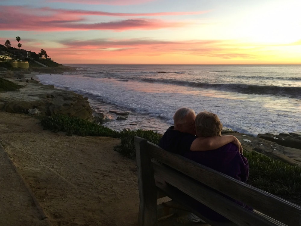 man and woman sitting on brown wooden bench in front of shoreline