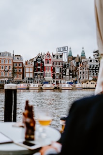 A picturesque view of a canal with traditional Dutch architecture in the background. Several canal boats are docked along the waterway, with buildings featuring ornate gables and a vibrant array of colors. In the foreground, a table with a bottle and a glass of beer creates a relaxed and inviting atmosphere.