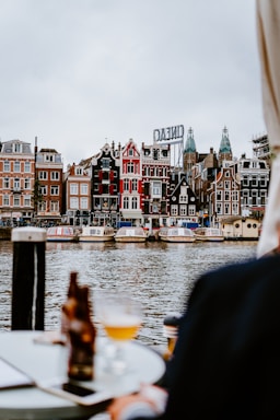 A picturesque view of a canal with traditional Dutch architecture in the background. Several canal boats are docked along the waterway, with buildings featuring ornate gables and a vibrant array of colors. In the foreground, a table with a bottle and a glass of beer creates a relaxed and inviting atmosphere.