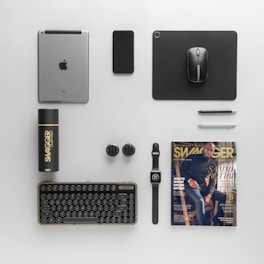 A neatly arranged flat lay featuring electronic devices, a magazine, a smartwatch, pens, a deodorant spray, and USB adapters. The items are organized on a light grey surface, including a keyboard, a black mouse on a mouse pad, an iPad, and a smartphone. There is also a magazine in the bottom right corner featuring a man on the cover swathed in warm tones.