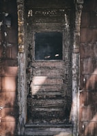 A weathered wooden door with peeling paint and a single small window in its upper half, surrounded by aged, textured wooden panels. The door has a rustic appearance, with dark, earthy colors and visible signs of decay.