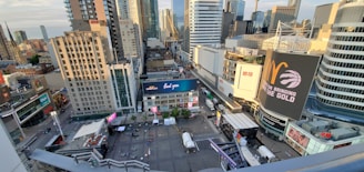 A bustling urban scene with tall modern skyscrapers under a blue sky. Numerous buildings are adorned with billboards and advertisements, while a plaza below has tents and seating areas. The scene suggests a vibrant city life.