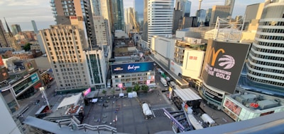 A bustling urban scene with tall modern skyscrapers under a blue sky. Numerous buildings are adorned with billboards and advertisements, while a plaza below has tents and seating areas. The scene suggests a vibrant city life.
