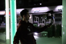 A person is holding a camera with a microphone attached, likely vlogging or recording. They appear to be outside, possibly at night or in a dimly lit area, with green and white illuminated columns nearby.