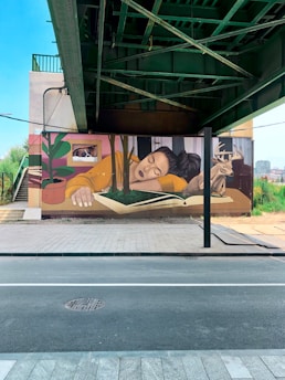 A mural painted on a wall underneath a bridge features a person with long dark hair wearing a yellow shirt, sleeping with their head resting on a book that has trees growing out of it. Nearby, a deer stands on the book, and there is a large potted plant to the left. Above the sleeping person, a cat peeks out from a window.