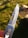 A close-up of a metallic folding knife blade with a sharp edge and a small hole near the top. The handle has two visible torx screws and is partially covered by a finger holding it. The background is a grassy and rocky area, slightly out of focus, in natural daylight.