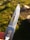 A close-up of a metallic folding knife blade with a sharp edge and a small hole near the top. The handle has two visible torx screws and is partially covered by a finger holding it. The background is a grassy and rocky area, slightly out of focus, in natural daylight.
