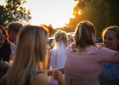 A group of people gathered outdoors, with a woman in a white dress and veil suggesting a wedding or special event. The setting sun casts a warm glow over the scene, creating a softly lit background with trees and blurred figures.