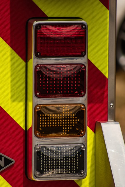 A close-up view of a vertical row of four rectangular lights on a vehicle, with colors red, amber, and white. The lights are set against a background with yellow and red diagonal stripes, resembling emergency vehicle markings. There's a metallic frame surrounding the lights, adding a reflective surface.