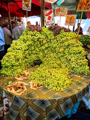 A market stall is covered with an abundant display of green and some red grapes arranged on a table with a colorful checkered tablecloth adorned with rose patterns. Several signs with Arabic writing and images hang above the grapes. In the background, people are seen browsing other produce under umbrellas.