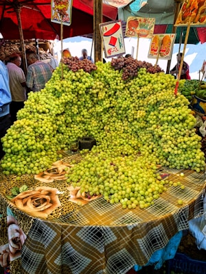 A market stall is covered with an abundant display of green and some red grapes arranged on a table with a colorful checkered tablecloth adorned with rose patterns. Several signs with Arabic writing and images hang above the grapes. In the background, people are seen browsing other produce under umbrellas.