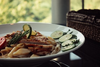 A white plate filled with creamy pasta topped with grilled chicken slices, garnished with fresh rosemary and lemon slices. The meal appears appetizing and is set on a dark table with natural light coming from a window, creating a cozy atmosphere. In the background, there is a woven basket and a blur of greenery outside.