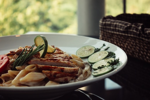 A white plate filled with creamy pasta topped with grilled chicken slices, garnished with fresh rosemary and lemon slices. The meal appears appetizing and is set on a dark table with natural light coming from a window, creating a cozy atmosphere. In the background, there is a woven basket and a blur of greenery outside.
