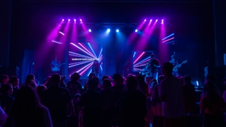 A concert scene with a band performing on a stage illuminated by vibrant purple and blue lights. A crowd gathers in front of the stage, and a digital screen behind the band displays colorful neon shapes and text.