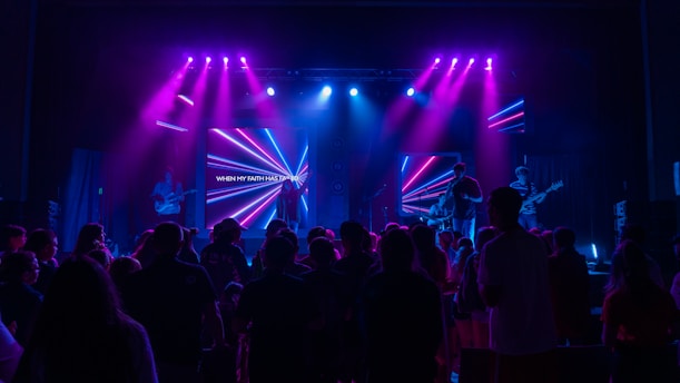 A concert scene with a band performing on a stage illuminated by vibrant purple and blue lights. A crowd gathers in front of the stage, and a digital screen behind the band displays colorful neon shapes and text.