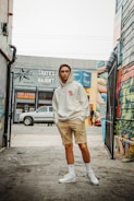 A young man is standing in an urban alleyway with graffiti-covered walls. He is wearing a light-colored hoodie, beige shorts, white socks, and white sneakers. There is an assortment of buildings and signs in the background, including a 'FOR LEASE' sign and a 'SKATES ON HAIGHT' sign. A pickup truck is parked on the street behind him.