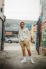 A young man is standing in an urban alleyway with graffiti-covered walls. He is wearing a light-colored hoodie, beige shorts, white socks, and white sneakers. There is an assortment of buildings and signs in the background, including a 'FOR LEASE' sign and a 'SKATES ON HAIGHT' sign. A pickup truck is parked on the street behind him.
