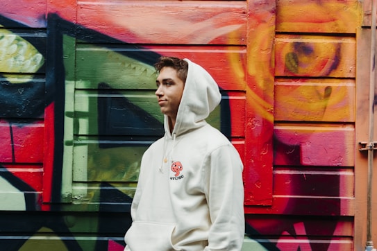 A person wearing a white hoodie with a red and black emblem stands against a vibrant, graffiti-covered wall. The wall features abstract patterns in red, green, yellow, and orange colors.