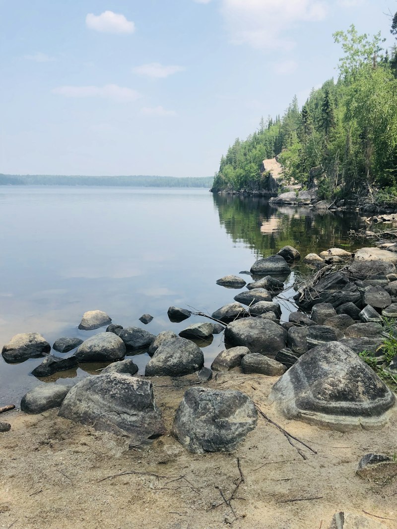 Rounded grey boulders on a sandy shoreline with a spruce-covered rocky point and a calm lake stretching to a far treeline