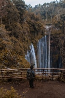 A photographer stands at a wooden barrier overlooking a large waterfall cascading down a rocky cliff surrounded by dense forest. The scene is lush with greenery, and the waterfall creates a misty atmosphere. The person appears to be setting up a camera on a tripod to capture the natural beauty.