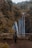 A photographer stands at a wooden barrier overlooking a large waterfall cascading down a rocky cliff surrounded by dense forest. The scene is lush with greenery, and the waterfall creates a misty atmosphere. The person appears to be setting up a camera on a tripod to capture the natural beauty.
