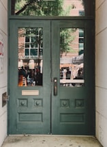 A pair of green wooden doors with decorative panels and two vertical windows. Outside, a reflection reveals a busy street scene with people, a tree, and a brick building with large windows.