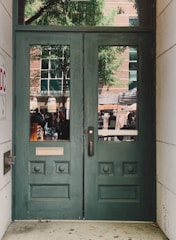 A pair of green wooden doors with decorative panels and two vertical windows. Outside, a reflection reveals a busy street scene with people, a tree, and a brick building with large windows.