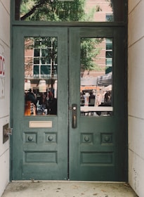 A pair of green wooden doors with decorative panels and two vertical windows. Outside, a reflection reveals a busy street scene with people, a tree, and a brick building with large windows.