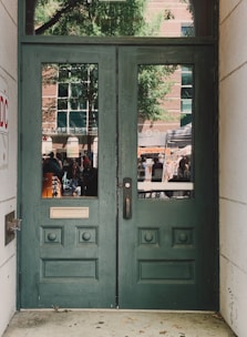A pair of green wooden doors with decorative panels and two vertical windows. Outside, a reflection reveals a busy street scene with people, a tree, and a brick building with large windows.
