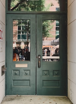 A pair of green wooden doors with decorative panels and two vertical windows. Outside, a reflection reveals a busy street scene with people, a tree, and a brick building with large windows.