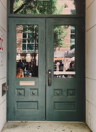 A pair of green wooden doors with decorative panels and two vertical windows. Outside, a reflection reveals a busy street scene with people, a tree, and a brick building with large windows.