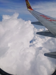 An airplane wing with a logo and text in red is visible above a dense layer of white clouds. The sky is clear with a gradient of blue near the top.