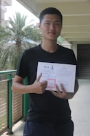 A person is standing in a corridor holding a diploma or certificate. They are wearing a black shirt and are giving a thumbs-up gesture. The background includes a palm tree and part of the building structure.