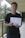 A person is standing in a corridor holding a diploma or certificate. They are wearing a black shirt and are giving a thumbs-up gesture. The background includes a palm tree and part of the building structure.