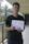A person is standing in a corridor holding a diploma or certificate. They are wearing a black shirt and are giving a thumbs-up gesture. The background includes a palm tree and part of the building structure.